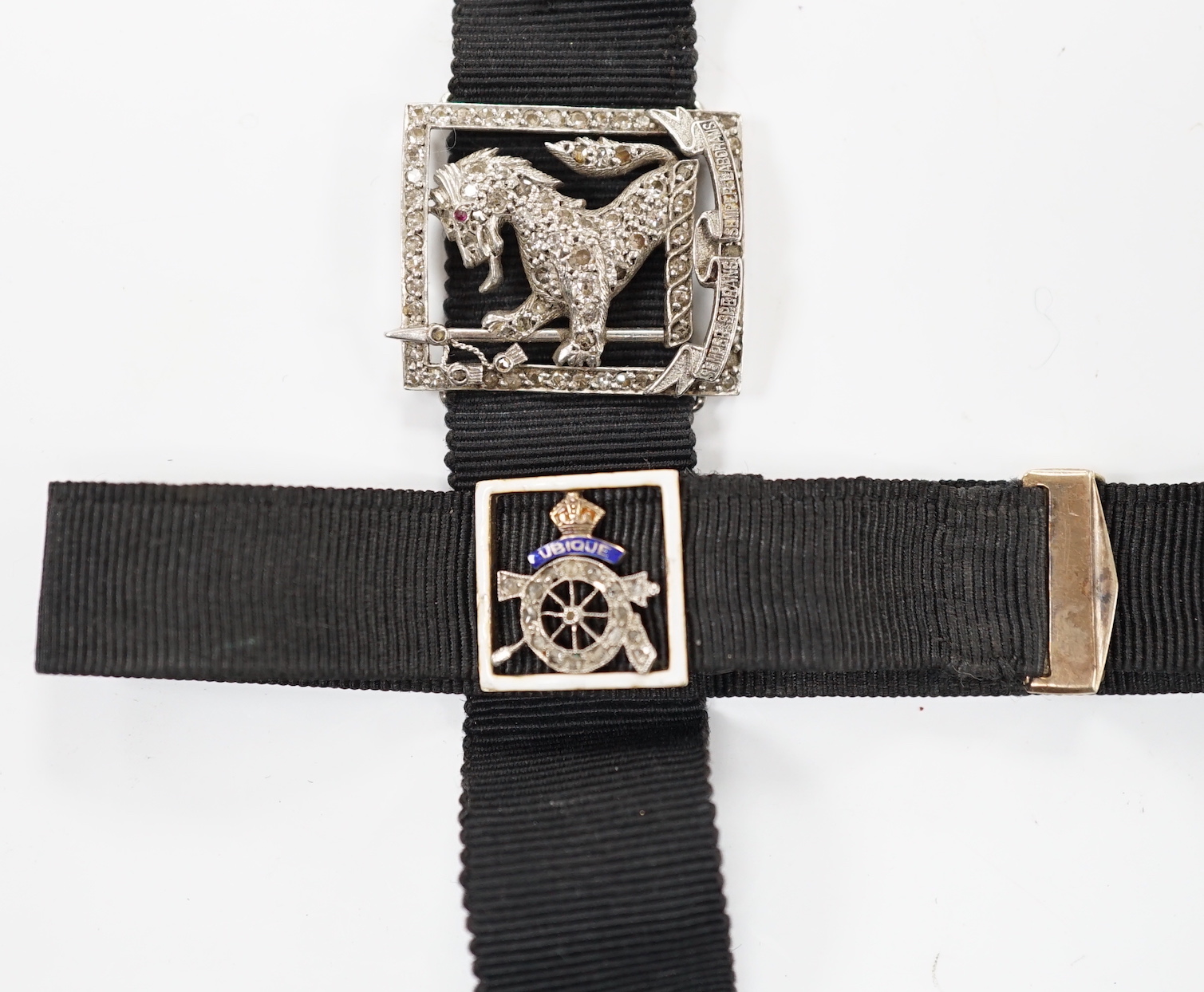 A black sash bracelet with diamond and enamel set Royal Fusiliers buckle, together with a similar sash with diamond set 'Charles' family crest, depicting a demi-rampant wolf, with motto.